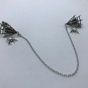 Może przedstawiać: Srebrny naszyjnik z łańcuszkiem z dwoma zawieszkami w kształcie pajęczyny i pająka. Zawieszki są przymocowane do łańcuszka srebrną pętelką.