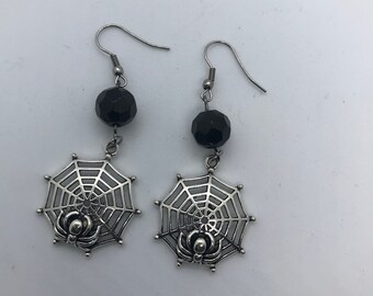 May include: A pair of silver spider web earrings with black beads. Each earring features a silver spider web with a spider in the center and a black bead hanging from the top.