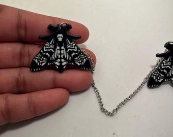 Könnte beinhalten: Schwarz-weiße Mottenbrosche mit Totenkopf-Design. Die Brosche ist durch eine silberne Kette mit einer zweiten Motte verbunden. Die Flügel der Motte weisen detaillierte Muster auf.