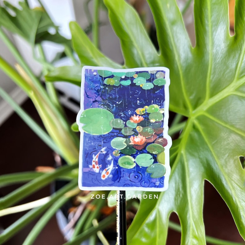 A rectangular sticker with a white border, showing two koi fish in a pond with lily pads and water lilies, is held up against a green plant.