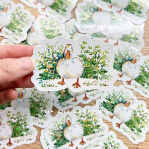 A hand holds a white vinyl sticker with a white duck in green foliage, surrounded by many identical stickers scattered on a surface, with a watermark at the bottom.