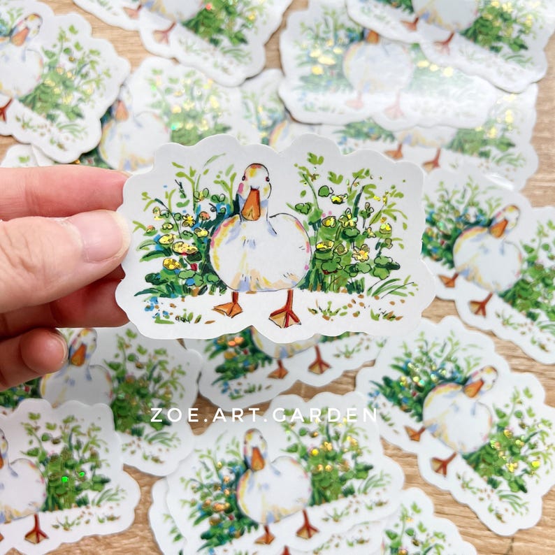 A hand holds a white vinyl sticker with a white duck in green foliage, surrounded by many identical stickers scattered on a surface, with a watermark at the bottom.