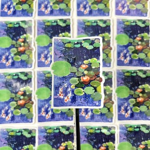 A rectangular sticker with a white border, showing two koi fish in a pond with lily pads and water lilies, is propped up by a pencil against a background of many identical stickers.