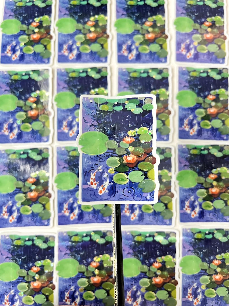 A rectangular sticker with a white border, showing two koi fish in a pond with lily pads and water lilies, is propped up by a pencil against a background of many identical stickers.