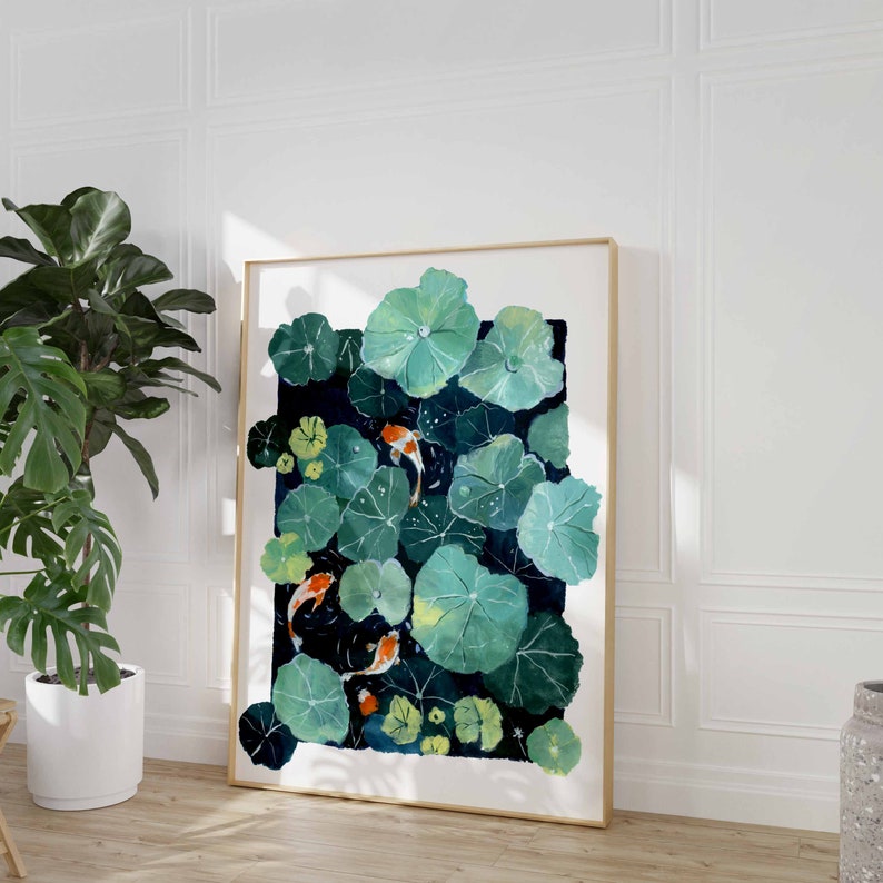 May include: A framed watercolor painting depicts a pond scene with koi fish swimming among lily pads. The artwork features shades of green, blue, and orange, with a gold frame. The painting is displayed on a wooden floor.