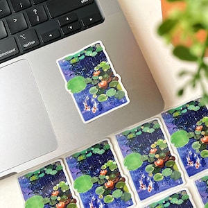 A rectangular sticker with a white border, depicting two koi fish in a pond with lily pads and water lilies, rests on a silver laptop keyboard.