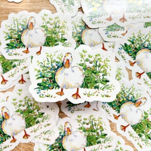 A white vinyl sticker with a white duck in green foliage is centered among many identical stickers on a light wooden surface, with a watermark at the bottom.