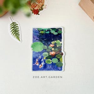 A rectangular sticker with a white border, depicting two koi fish in a pond with lily pads and water lilies, is centered on a white surface with green leaves and a small house sticker nearby.