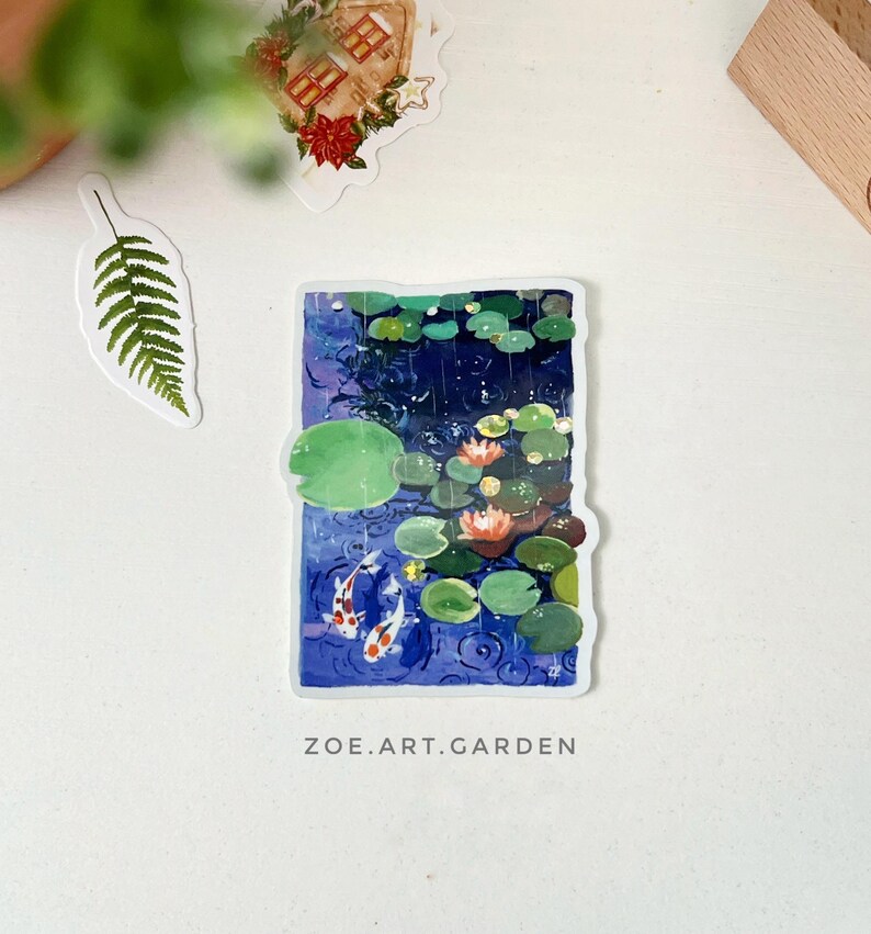 A rectangular sticker with a white border, depicting two koi fish in a pond with lily pads and water lilies, is centered on a white surface with green leaves and a small house sticker nearby.