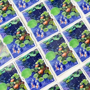 An overhead view of many rectangular stickers with white borders, each showing two koi fish in a pond with lily pads and water lilies, arranged in a grid pattern.