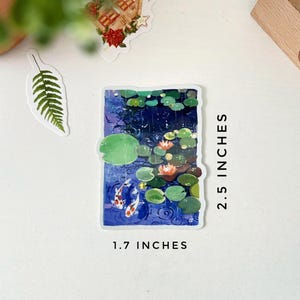 A rectangular sticker with a white border, showing two koi fish in a pond with lily pads and water lilies, is displayed next to text indicating its dimensions: "2.5 INCHES" and "1.7 INCHES."