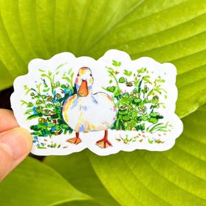 A hand holds a white vinyl sticker depicting a white duck with an orange beak and feet, standing in green foliage with small flowers, against a bright green leaf background.
