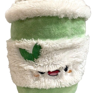 May include: A plush green and white stuffed toy shaped like a cup of matcha. The cup has a smiling face with embroidered details, a white fluffy rim, and a green base. Two green leaves are on the front.