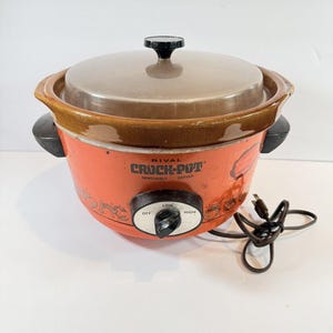 May include: An orange Rival Crock-Pot slow cooker with a brown ceramic rim and a clear glass lid. The front features a black control knob with Off, Low, and High settings, along with black side handles, a power cord, and printed food graphics.