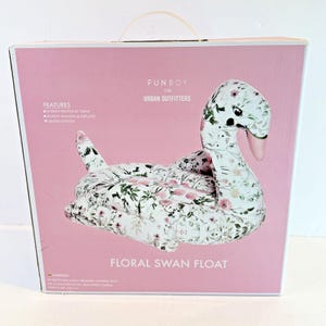 May include: A square pink product box for a Funboy for Urban Outfitters floral swan float. The box features a graphic of a white inflatable swan with a pink and green floral pattern, along with text listing product features and warnings.
