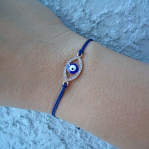 Op de afbeelding: Een blauwe koordarmband met een goudkleurige, oogvormige bedel. De bedel is bezet met heldere kristallen en een blauw en wit oogontwerp. De armband wordt om de pols gedragen.