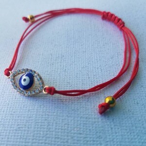 Op de afbeelding: Rode armband met een goudkleurige evil eye bedel met een blauw en wit centrum, omringd door heldere kristallen. De armband heeft verstelbare rode koorden en goudkleurige kralen.