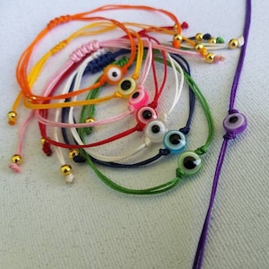 May include: A set of colorful string bracelets with evil eye charms. The bracelets are made of different colors of string, including orange, pink, yellow, red, white, blue, and green. The evil eye charms are all different colors, including blue, green, pink, and purple.