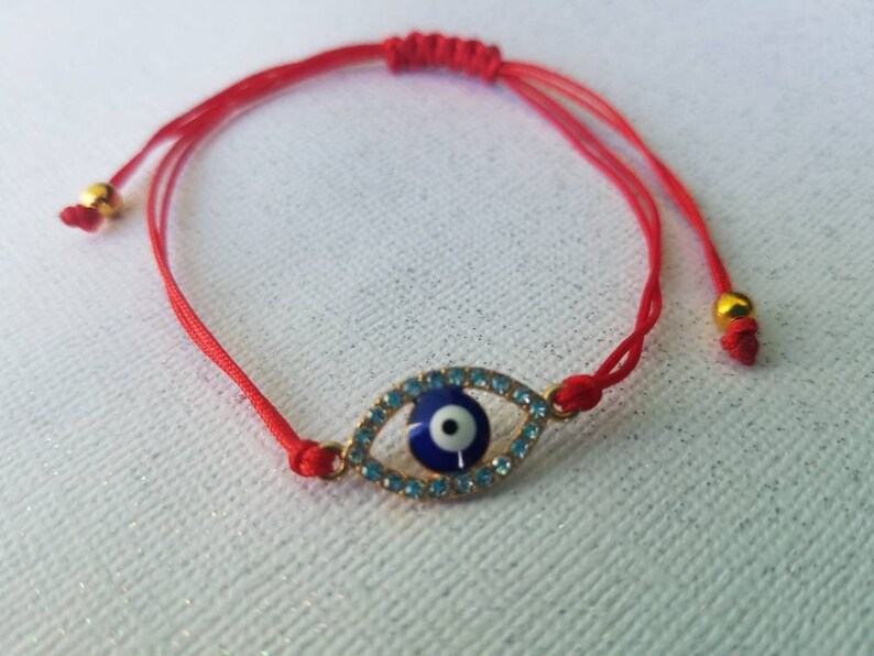Op de afbeelding: Een rode koordarmband met een goudkleurige evil eye bedel. De bedel heeft een blauw centrum met een witte pupil, omringd door blauwe kristallen. De armband is verstelbaar met schuifknopen en goudkleurige kralen.