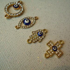 Op de afbeelding: Vier goudkleurige bedels met heldere kristallen accenten en blauw-witte evil eye-ontwerpen. De bedels omvatten een cirkel, een oogvorm, een hand en een kruis. Elke bedel heeft kleine lussen om aan sieraden te bevestigen.