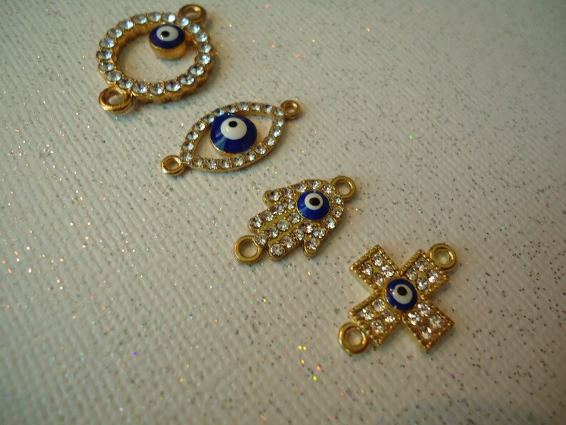 Op de afbeelding: Vier goudkleurige bedels met heldere kristallen accenten en blauw-witte evil eye-ontwerpen. De bedels omvatten een cirkel, een oogvorm, een hand en een kruis. Elke bedel heeft kleine lussen om aan sieraden te bevestigen.