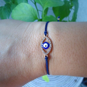 Op de afbeelding: Een marineblauwe koordarmband met een goudkleurige, ovale bedel. De bedel heeft een blauw oogontwerp en is bezet met kleine, heldere kristallen. Deze delicate armband is een sieraad.