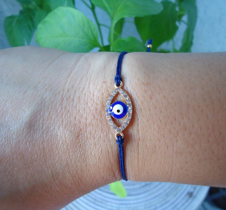 Op de afbeelding: Een marineblauwe koordarmband met een goudkleurige, ovale bedel. De bedel heeft een blauw oogontwerp en is bezet met kleine, heldere kristallen. Deze delicate armband is een sieraad.