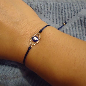 Op de afbeelding: Marineblau touw armband met een goudkleurige, oogvormige bedel. De bedel is bezet met kleine, heldere kristallen en een blauw en wit oogontwerp. De armband heeft een verstelbare schuifknoop.
