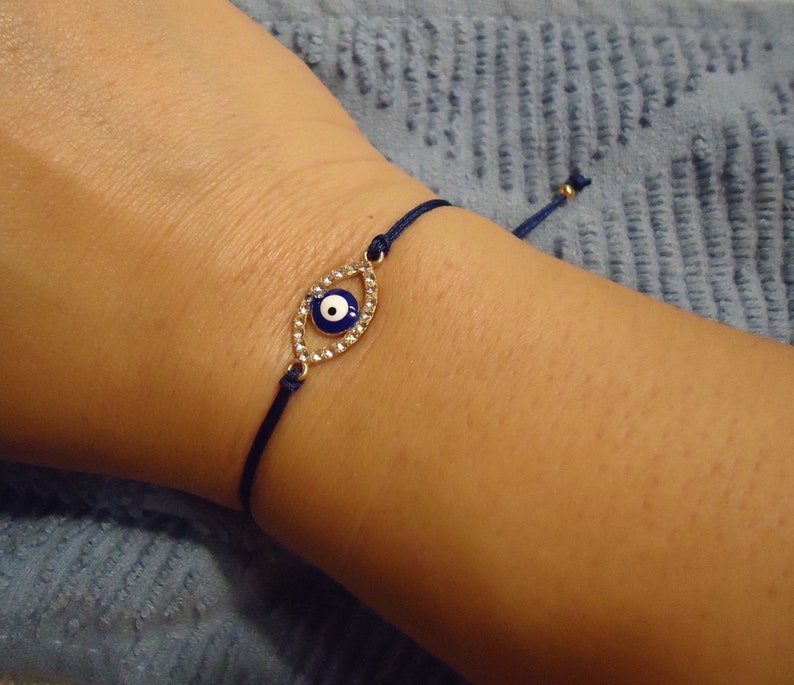 Op de afbeelding: Marineblau touw armband met een goudkleurige, oogvormige bedel. De bedel is bezet met kleine, heldere kristallen en een blauw en wit oogontwerp. De armband heeft een verstelbare schuifknoop.