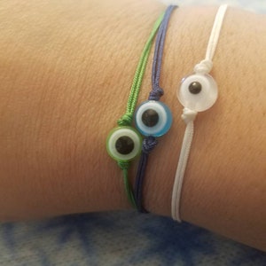 May include: Three string bracelets with evil eye charms. The charms are blue, green, and white. The bracelets are tied on a person's wrist.