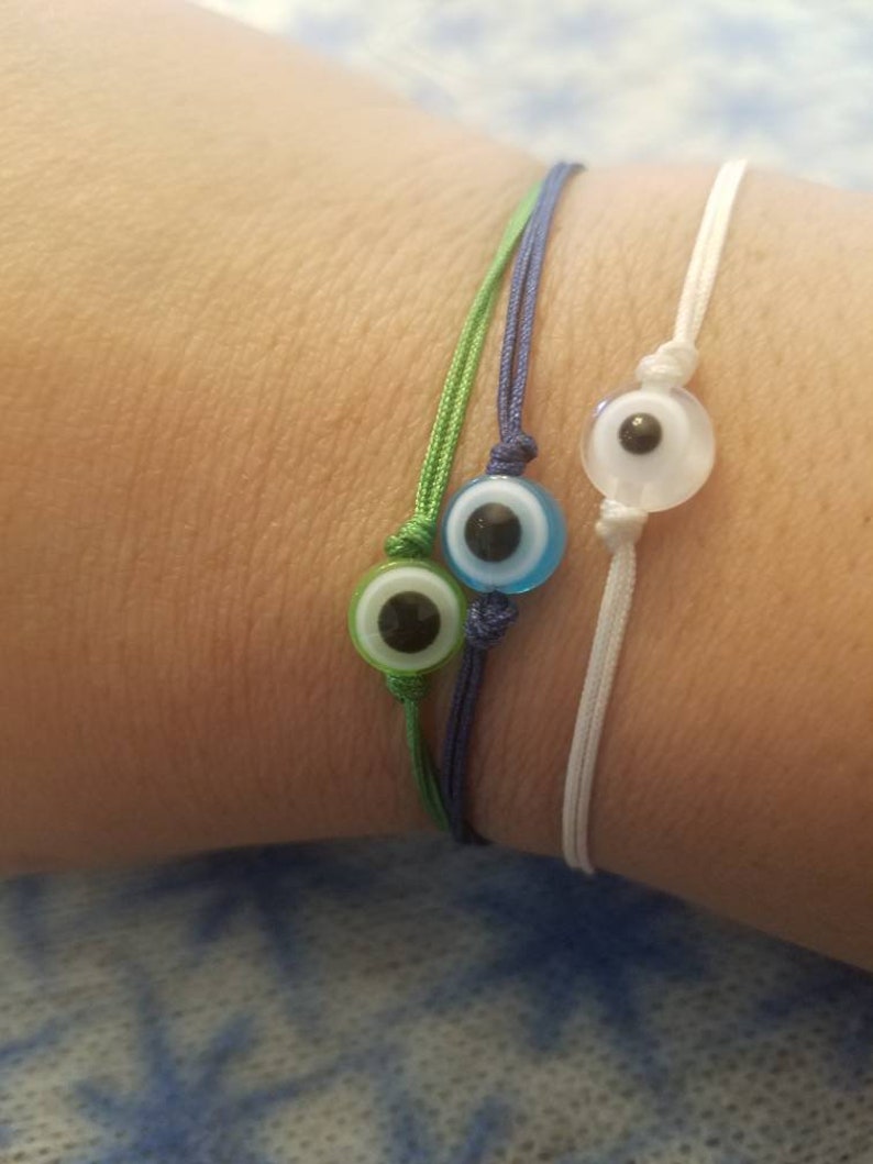 May include: Three string bracelets with evil eye charms. The charms are blue, green, and white. The bracelets are tied on a person's wrist.