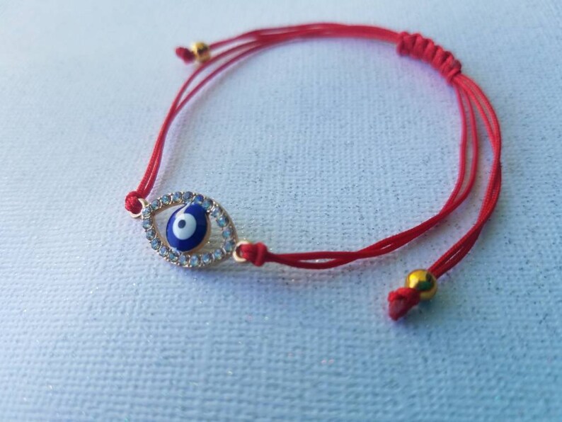 Op de afbeelding: Rode armband met een goudkleurige evil eye bedel met een blauw en wit centrum, omringd door heldere kristallen. De armband heeft verstelbare rode koorden en goudkleurige kralen.