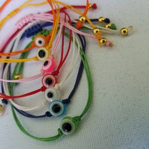 May include: A set of colorful string bracelets with evil eye charms. The bracelets are made of different colors of string, including red, green, blue, white, and yellow. Each bracelet has a gold bead at the end and a small evil eye charm in the center.