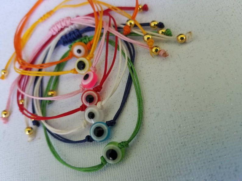 May include: A set of colorful string bracelets with evil eye charms. The bracelets are made of different colors of string, including red, green, blue, white, and yellow. Each bracelet has a gold bead at the end and a small evil eye charm in the center.