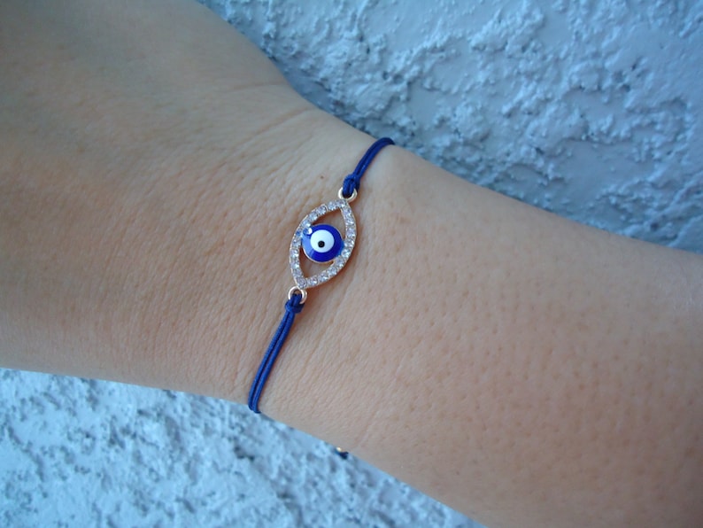 Op de afbeelding: Een blauwe koordarmband met een goudkleurige, oogvormige bedel. De bedel is bezet met heldere kristallen en een blauw en wit oogontwerp. De armband wordt om de pols gedragen.