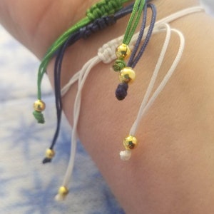 May include: A multi-colored friendship bracelet with green, blue, and white strings and gold beads.