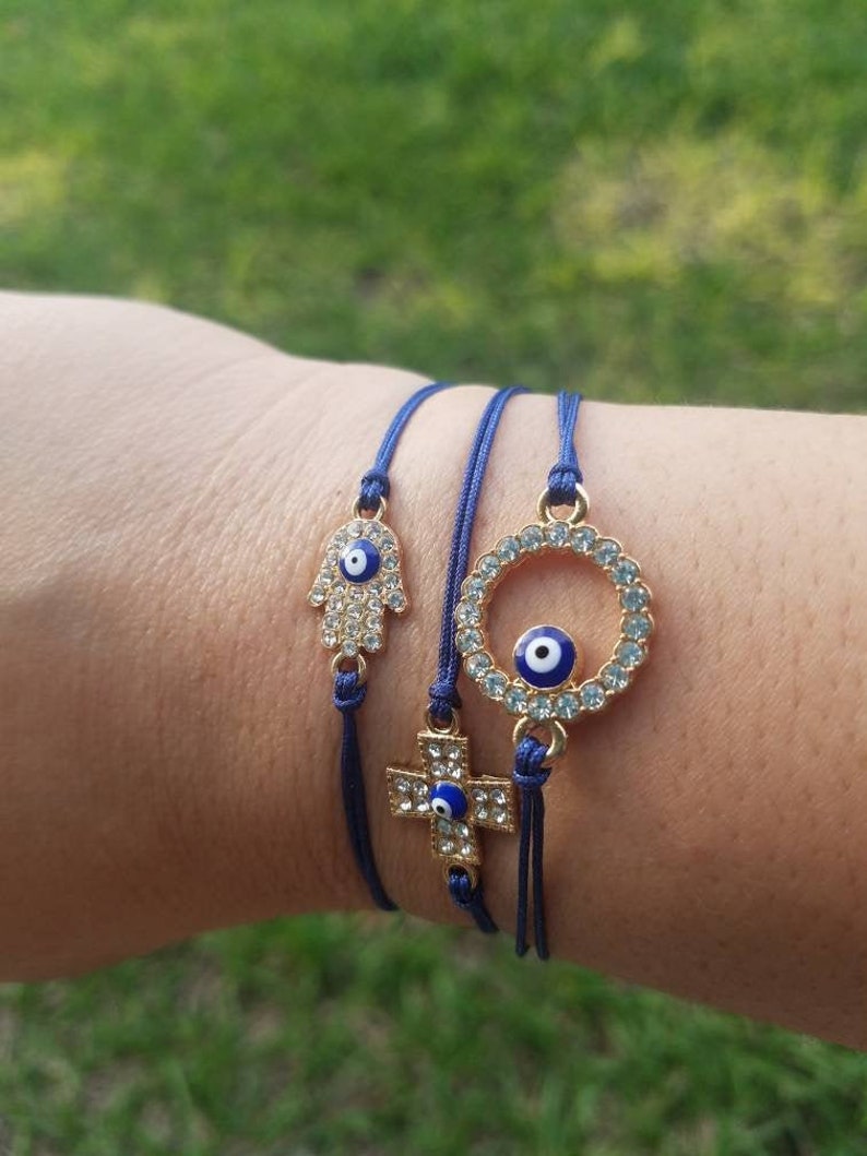 Op de afbeelding: Drie blauwe touwarmbanden met goudkleurige bedels. E&eacute;n armband heeft een Hamsa-hand, een andere een kruis en de derde een cirkel, allemaal versierd met heldere kristallen en een blauw oog. De armbanden worden getoond tegen een groene achtergrond.