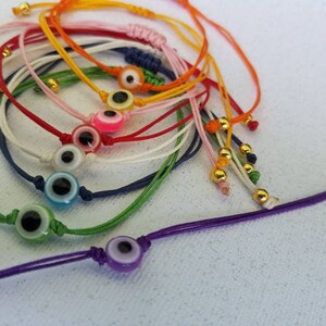 May include: A set of colorful string bracelets with evil eye charms. The bracelets are made of different colors of string, including red, blue, green, yellow, and white. Each bracelet has a small, round evil eye charm with a blue eye and a white pupil.