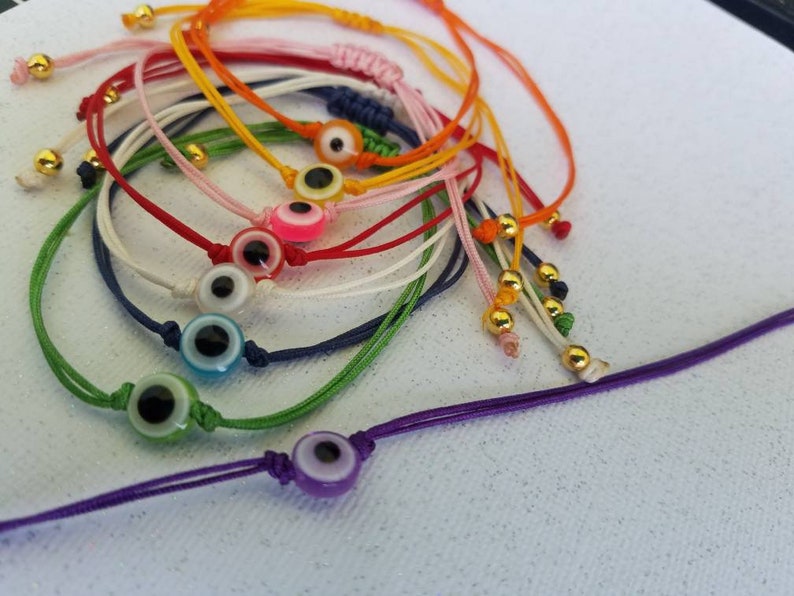 May include: A set of colorful string bracelets with evil eye charms. The bracelets are made of different colors of string, including red, blue, green, yellow, and white. Each bracelet has a small, round evil eye charm with a blue eye and a white pupil.