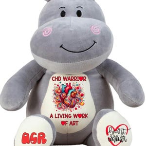Può includere: Ippopotamo di peluche grigio con un cuore rosso e il testo "CHD Warrior" e "A Living Work of Art" sul petto. L'ippopotamo ha una macchia bianca sul ventre con il testo "AGR" in rosso. L'ippopotamo ha una macchia bianca sul piede destro con il testo "heart warrior" in rosso.