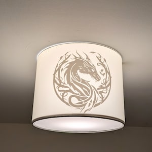 May include: A cream-coloured cylindrical lampshade, featuring a detailed dragon design in a lighter cream shade, encircled by stylized foliage. The lampshade is mounted on a ceiling fixture.