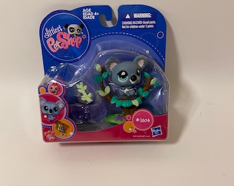 May include: A gray koala plush toy sitting on a green tree branch with blue leaves. The toy is part of the Littlest Pet Shop toy line and is packaged in a clear plastic box with a pink and blue background. The box also features a panda and the text "Littlest Pet Shop" and "#1604".