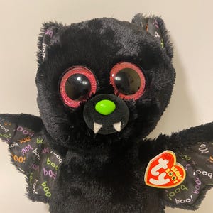 May include: Black plush bat with large pink and red eyes and a green nose. The bat has a pink mouth with white teeth and wings with a "boo" pattern. The bat has a Ty tag that says "Beanie Boo's".
