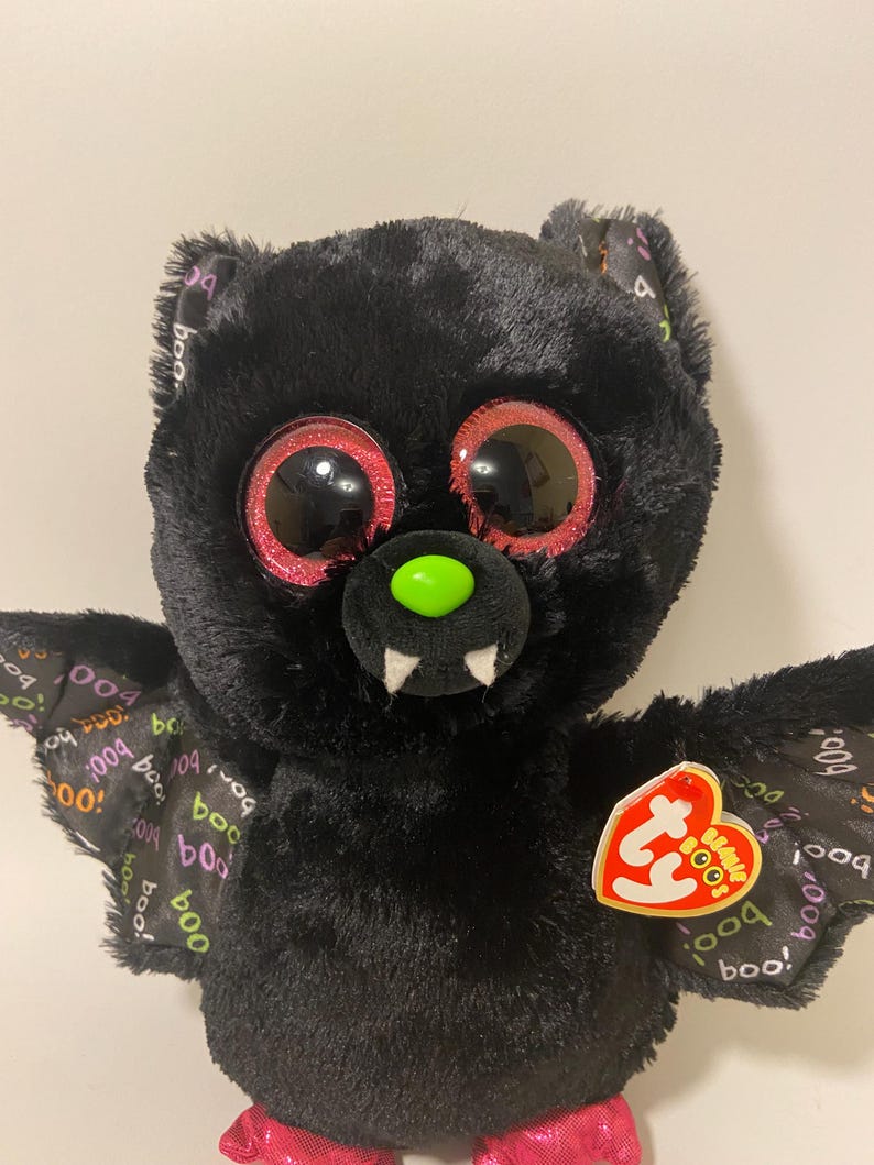 May include: Black plush bat with large pink and red eyes and a green nose. The bat has a pink mouth with white teeth and wings with a "boo" pattern. The bat has a Ty tag that says "Beanie Boo's".