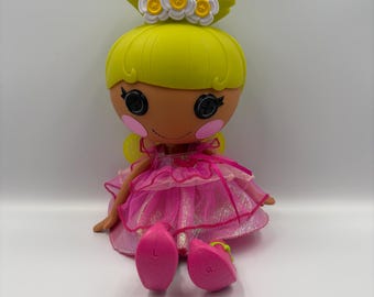 May include: A Lalaloopsy doll with a yellow head and hair, wearing a pink dress with a layered skirt. The doll has button eyes, rosy cheeks, and a flower crown. The doll's pink shoes are marked with an "L" and an "R".