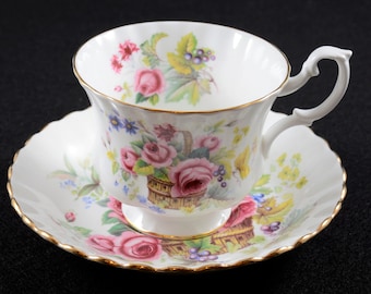 May include: A white teacup and saucer set with a floral pattern featuring pink roses, green leaves, and yellow flowers. The set has a gold trim around the edges.