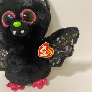 May include: Black plush bat with pink glitter eyes and a green nose. The bat has pink feet and a Ty Beanie Boo tag with the words "Boo!" and "004".