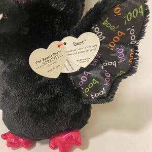 May include: Black plush bat with pink glitter eyes and pink feet. The bat has a tag that reads "The Beanie Boo's Collection" and "Dart". The tag also says "Sometimes I can act a little batty When I eat a peppermint patty!" and "My Birthday is October 18". The bat's wings have a black and white pattern with the word "boo" repeated.