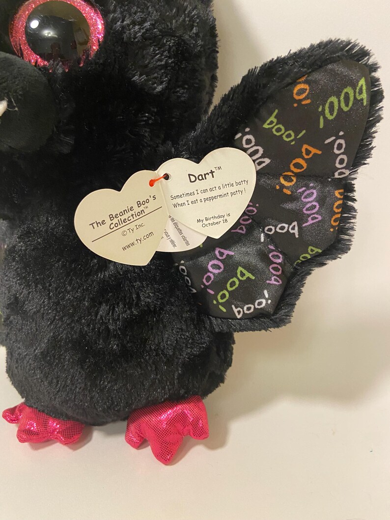 May include: Black plush bat with pink glitter eyes and pink feet. The bat has a tag that reads "The Beanie Boo's Collection" and "Dart". The tag also says "Sometimes I can act a little batty When I eat a peppermint patty!" and "My Birthday is October 18". The bat's wings have a black and white pattern with the word "boo" repeated.