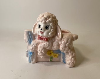 May include: A ceramic planter shaped like a pink lamb sitting on a blue fence with yellow flowers.